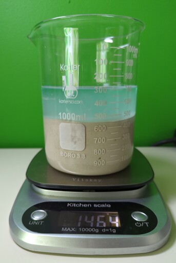 A beaker on a scale. The beaker is full of gray powder to the 450 mL line and blue tinted water to the 700 mL line. The scale reads 1464 grams.